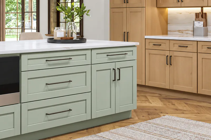 Sage green shaker island with dark bar pulls and a white countertop beside natural wood cabinets